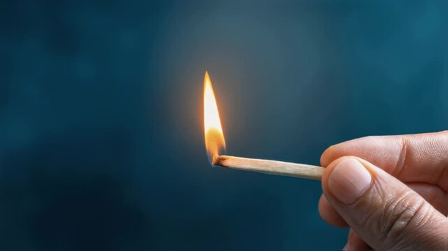 Hand holding wooden match with bright flame burning, creating contrast against deep teal background. Close-up of single match moment of lighting.