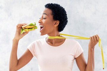 Cheat meal or diet breakdown concept. Black lady trying to eat unhealthy burger, pulling herself...