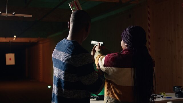 Shooting gallery staff member teaching student how to hold and handle handgun. Firing range expert providing guide to african american woman learning how to achieve proficiency with guns, camera A