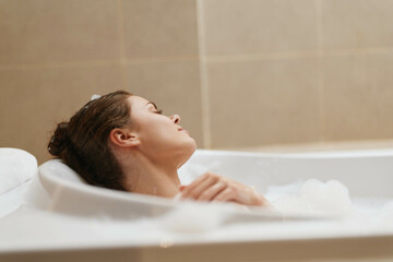 Relaxed young woman enjoying a bubble bath, eyes closed in tranquility, surrounded by soft foam and warm lighting. Self-care and wellness concept.