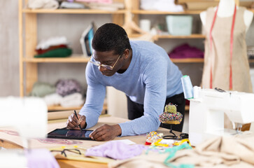 Fototapeta premium Dedicated male tailor concentrating on designing custom dress using tablet, reviewing and adjusting clothing model in well-equipped creative studio