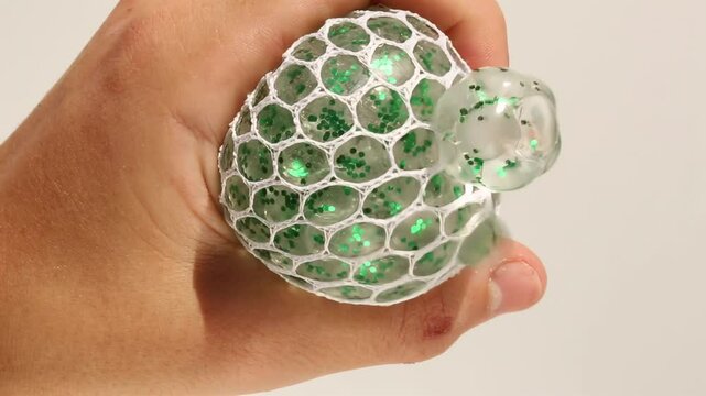 Hand holding transparent stress ball with green hydrogel beads inside. The fingers squeeze and rotate the clear ball showing the movement of the green orbs floating in the gel.
