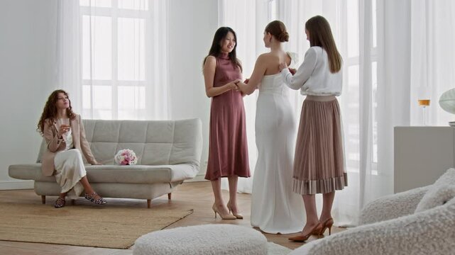 Two bridesmaids adjusting wedding dress and hairstyle of bride while third one resting on comfortable sofa with glass of champagne