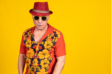 Stylish senior man wearing hawaiian shirt and fedora hat posing on yellow background
