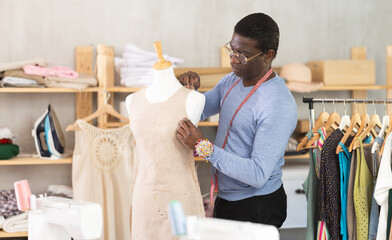 African American man tailor connects parts of dress on mannequin. Female craftsman attaches front...