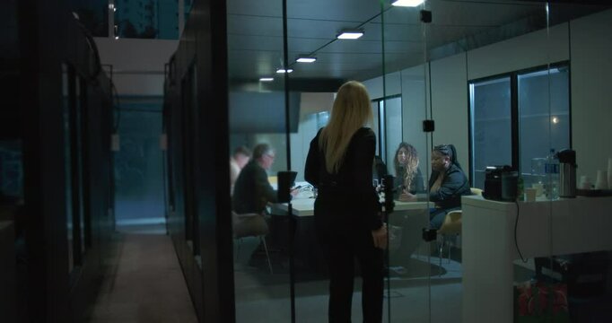 Female executive entering glass boardroom as colleagues sit around table with laptops during late office meeting in modern corporate workspace - Powered by Adobe