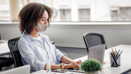 New normal and office work with protective mask and antiseptic. African american woman typing on...