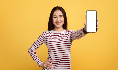 Naklejka premium Asian young woman with long dark hair is smiling while holding a smartphone in front of her. The vibrant yellow background enhances her cheerful demeanor and the blank screen invites curiosity.