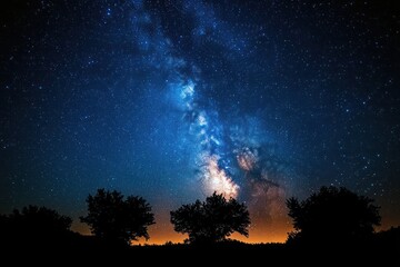 Fototapeta premium Clear night sky with bright Milky Way galaxy and stars over silhouette of trees at horizon glowing with warm light
