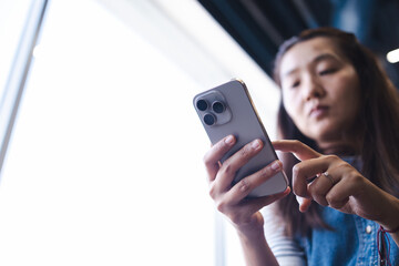 Fototapeta premium Asian woman engaging with ai technology through smartphone connection, focused on device in modern setting with natural light from window