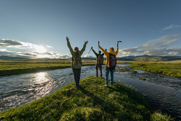 A group of people in bright clothes and with backpacks on their backs, stand with their hands raised in a green meadow during the golden hour. The green meadow is surrounded by rivers.