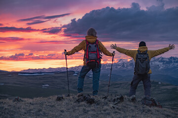 Two travelers in bright clothes with backpacks on their backs stand on the top of a mountain during a fiery sunset, overlooking the mountains that are partially covered with snow. One of them stands w
