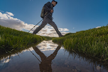 A traveler with a backpack on his back steps over a stream surrounded by green grass and is reflected in the water. Reflection of a person in water in nature. Trekking poles for traveling.