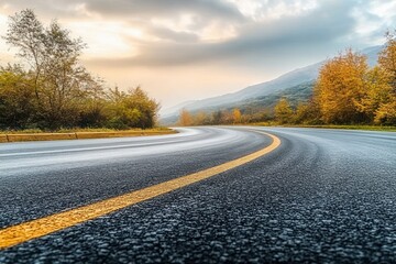 Fototapeta premium Empty curved asphalt road with yellow center line during autumn surrounded by trees with fall foliage and mountains under a cloudy sky at sunset