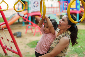 mother holding her daughter to hanging on playground rings in park.