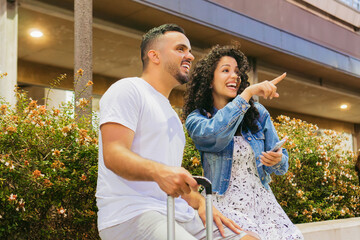Young Latin couple, tourists, sitting on a plaza bench with flowers, happily pointing at something they see