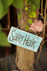 Blue wooden sign with the words “Salty Hair” hanging on a tree in a tropical setting. Whimsical beach decor and carefree summer vibes in a peaceful island garden.