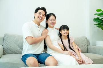 Asian family (father, daughter, and mother) sitting closely on a sofa and smiling at the camera.