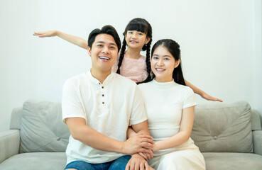 Asian family portrait. The daughter playfully spreads her arms like wings from behind her smiling parents on the sofa.