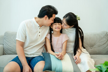 Fototapeta premium Asian parents kissing their happy daughter on both cheeks as they sit together on the sofa.