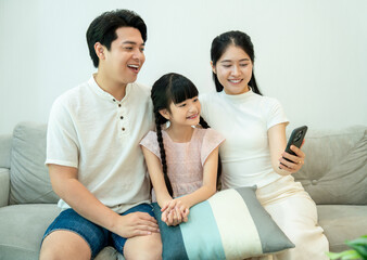Asian family sitting on the sofa, looking at a smartphone together, enjoying a video, movie, or a...