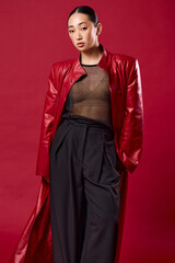 Fashion asian woman in red leather jacket and black pants posing against red background with hands on hips