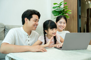 Obraz premium Asian family on a video call. The little daughter waves happily at the laptop screen while sitting with her parents.