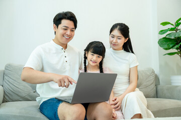 Fototapeta premium Asian father points at a laptop screen, teaching his daughter or showing her something online, with the mother looking on.