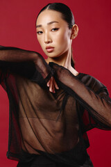 Fashion asian woman in sheer top and black pants poses against a red background with hands on hips and head tilted to the side