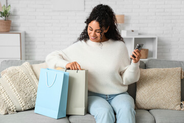 Happy young African-American woman with shopping bags and mobile phone at home