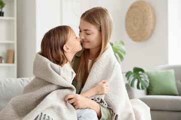 Cute little girl and her mother with warm plaid touching noses on sofa at home