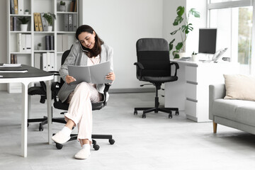 Young businesswoman with folder talking by mobile phone at table in office. National Wear Your...