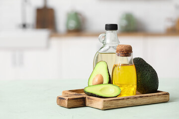 Bottles of fresh oil and wooden board with avocados on green table in kitchen