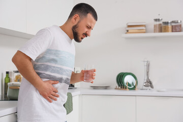 Young man with glass of water suffering from bellyache in kitchen