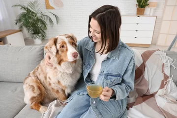 Zelfklevend Fotobehang Happy young woman with cute Australian Shepherd dog and cup of tea on sofa at home © Pixel-Shot