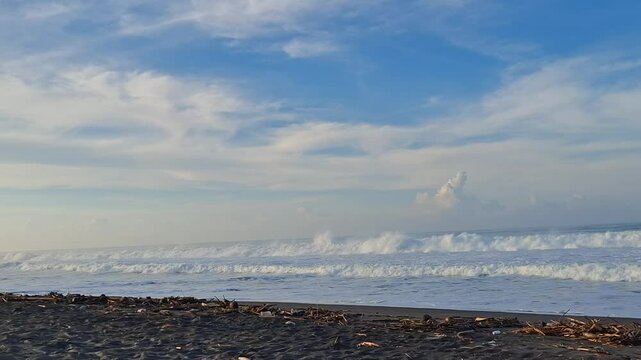 A wide view of a tranquil black sand beach with strong waves crashing under a bright blue sky. The scene evokes a peaceful yet powerful coastal vibe, perfect for themes of nature, travel, serenity, an