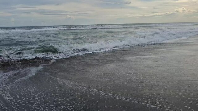 A close-up footage of gentle ocean waves rolling over wet sand, creating soft foam patterns as the tide flows in. The cloudy sky above adds a calm and moody coastal atmosphere. Ideal for nature themes