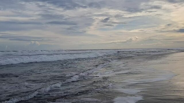 A wide footage of a peaceful tropical beach with gentle waves rolling onto the shore under an overcast sky. The ocean surface reflects soft evening light, creating a tranquil and cinematic coastal sce