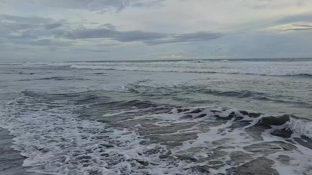 Peaceful view of gentle sea waves approaching the shore under a cloudy sky. White sea foam spreads over the wet sand, creating a calming and natural atmosphere
