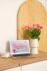 Laptop with text HAPPY WOMEN'S DAY, cup and tulips on chest of drawers in room