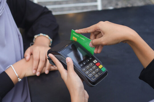 Credit card payment using a point of sale terminal. Close-up of hand swiping a green card while another person waits at a table. Modern cashless transaction concept.
