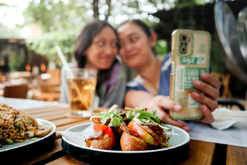 Young Asian mother and her teenage daughter smiling and taking selfie together using smartphone while having lunch at an outdoor restaurant. Close family relationship concept.