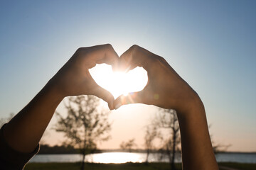 Female hands making heart gesture against river at sunset. Summer solstice