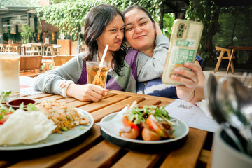Young Asian mother and her teenage daughter smiling and taking selfie together using smartphone while having lunch at an outdoor restaurant. Close family relationship concept.