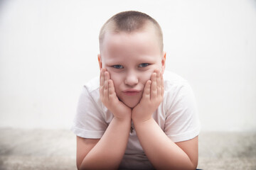Boy with short hair sits with hands on cheeks, displaying a pouting expression in a light-colored...
