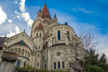 Naklejka premium Stunning view of St. Francis of Assisi Church in Vienna, Austria, showcasing its Romanesque Revival architecture, red rooftops, and majestic towers under a vivid blue sky with scattered clouds.