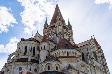 Fototapeta premium Magnificent view of St. Francis of Assisi Church in Vienna, Austria, featuring its iconic Romanesque Revival architecture, clock towers, and red-tiled rooftops against a bright sky.