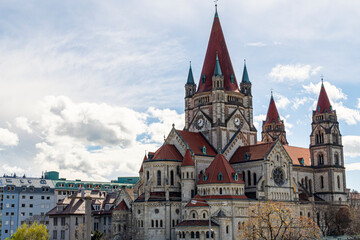 Fototapeta premium Stunning view of St. Francis of Assisi Church in Vienna, Austria, showcasing Romanesque Revival architecture with red rooftops, multiple spires, and a bright sky backdrop.