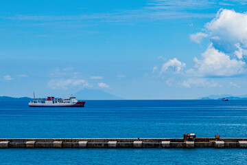 Large ferry ship sailing on calm blue ocean with mountain and clouds in the background, viewed from a concrete pier on a clear sunny day.