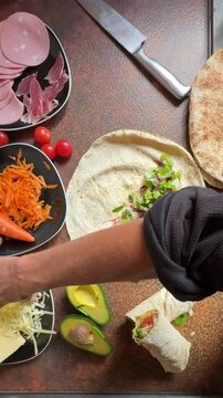 overhead view, cropped female hands preparing healthy food, making sandwich wrap with lettuce, grated cheese, grated carrots and mortadella at home in kitchen